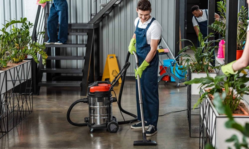 young-cleaner-cleaning-floor-with-vacuum-cleaner-near-multicultural-colleagues.jpg